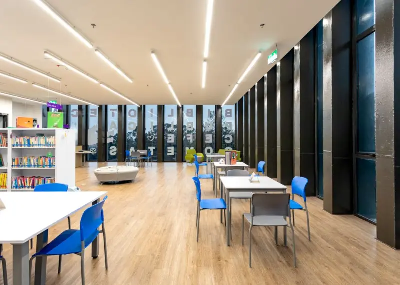 Biblioteca juvenil, con mesas de madera y sillas azúles de plástico.